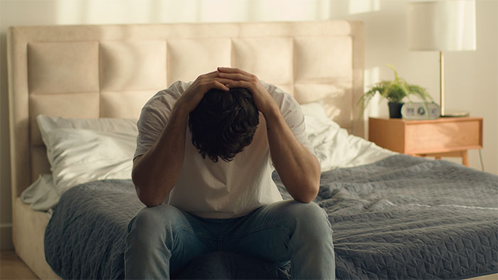 Man sitting on edge of bed with head in hands, appearing distressed after girlfriend kisses another woman. Man sitting on edge of bed with head in hands, appearing distressed after girlfriend kisses another woman.