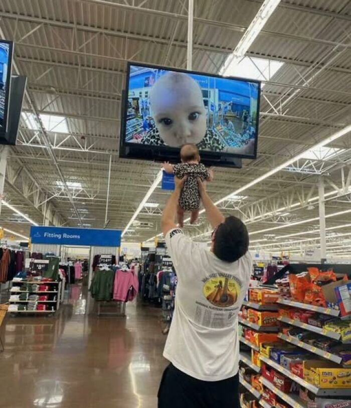 Man lifting baby in store aisle with baby’s face displayed on overhead security screen, a popular pic that goes hard