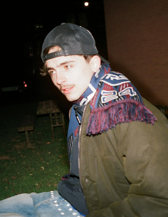Young man wearing a backward cap and a patterned scarf sitting outdoors at night, linked to Timothée Chalamet and EsDeeKid theory. Young man wearing a backward cap and a patterned scarf sitting outdoors at night, linked to Timothée Chalamet and EsDeeKid theory.