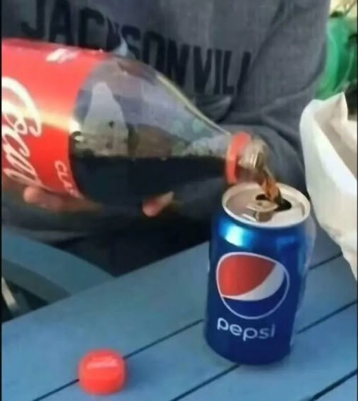 Person pouring Coca-Cola from a bottle into a Pepsi can on a blue table in a casual setting, showing pics that go hard.