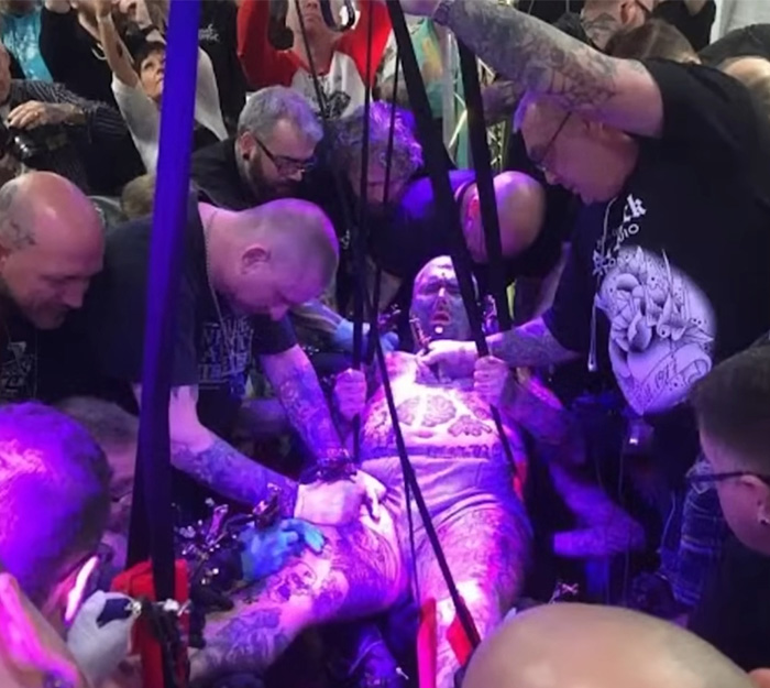 Man covered in tattoos getting eyeball tattooed by a group of tattoo artists during a ritual with purple lighting. Man covered in tattoos getting eyeball tattooed by a group of tattoo artists during a ritual with purple lighting.