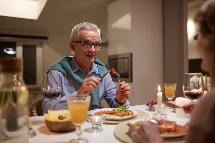 Elderly man in glasses discussing Thanksgiving arguments while eating dinner with wine and candles at a cozy table setting.