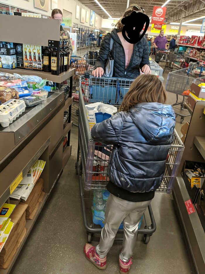 Child leaning on shopping cart while waiting in line behind shopper inside grocery store, faith in humanity restored moment.