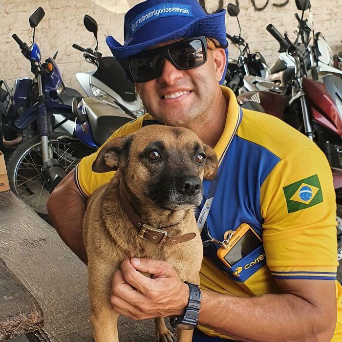 Mailman in Brazil wearing uniform and hat, smiling while holding a brown dog, surrounded by motorcycles in the background.