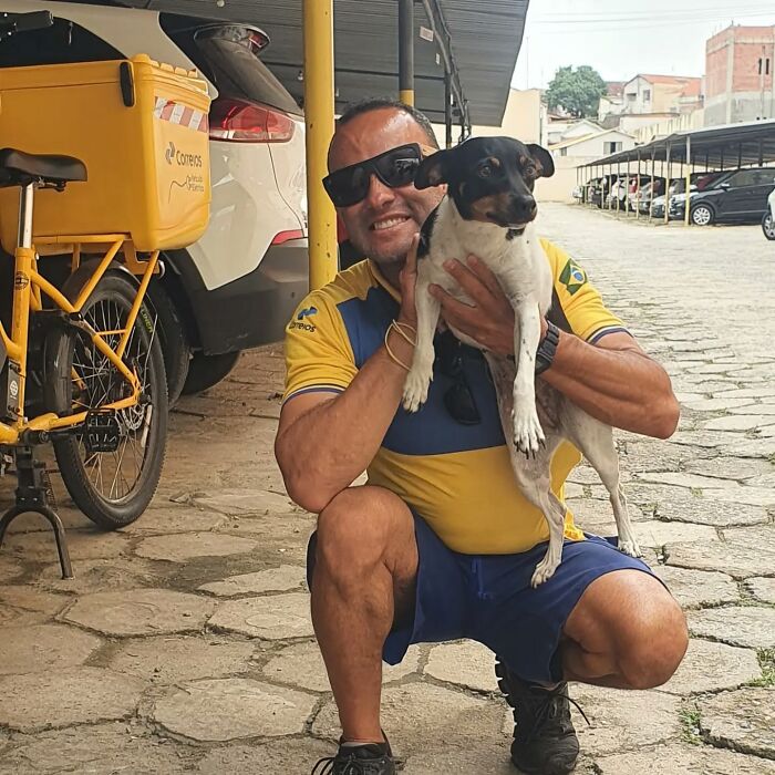 Mailman wearing sunglasses holding a small dog on a street near his yellow postal bicycle during his delivery route.