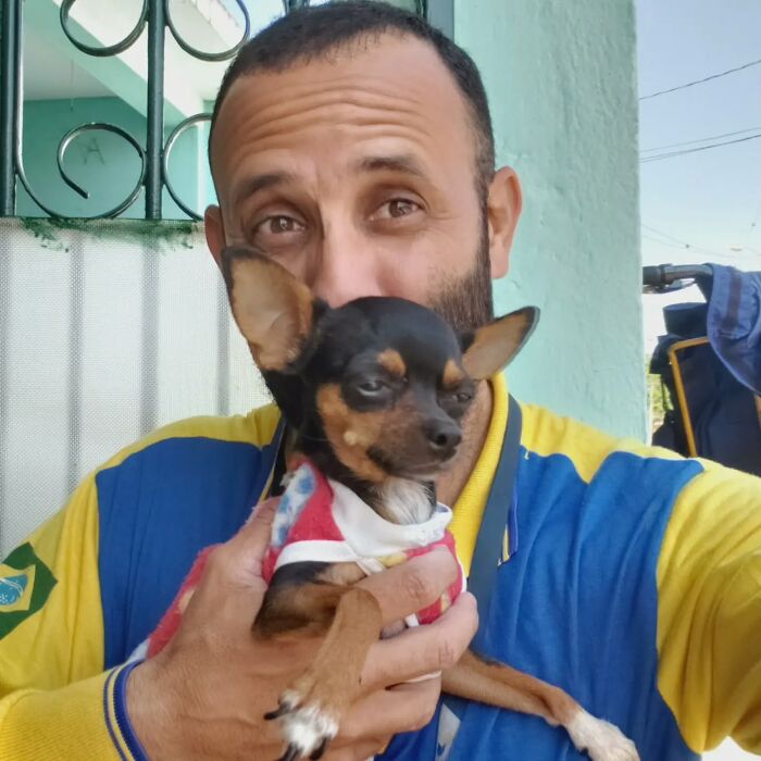 Mailman wearing a blue and yellow uniform happily holding a small dog, showing the bond between the mailman and dogs on his route.