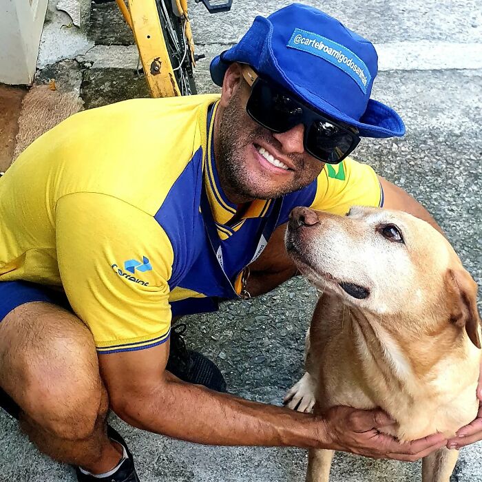 Mailman wearing blue cap and sunglasses smiling while petting a friendly dog on his delivery route outdoors.