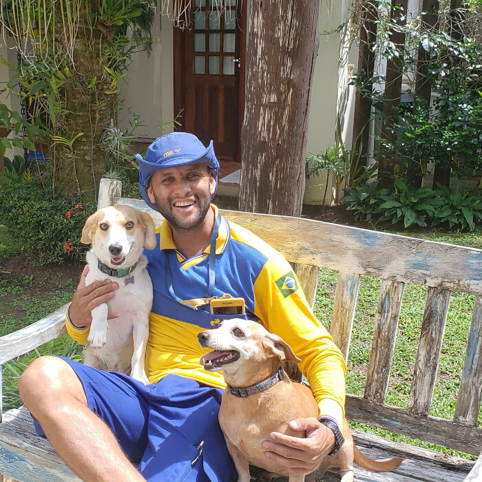Mailman in yellow and blue uniform sitting on a bench, happily holding two friendly dogs on his delivery route.
