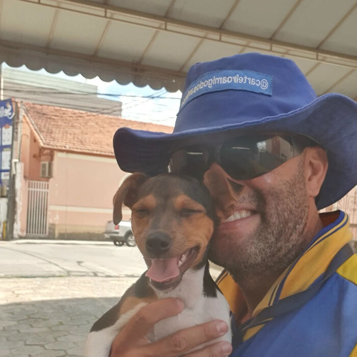 Mailman wearing blue hat and sunglasses smiling while holding a happy small dog on his delivery route outdoors.
