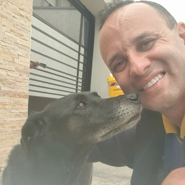 Mailman smiling while a black dog affectionately nuzzles his face during a friendly moment on his route.