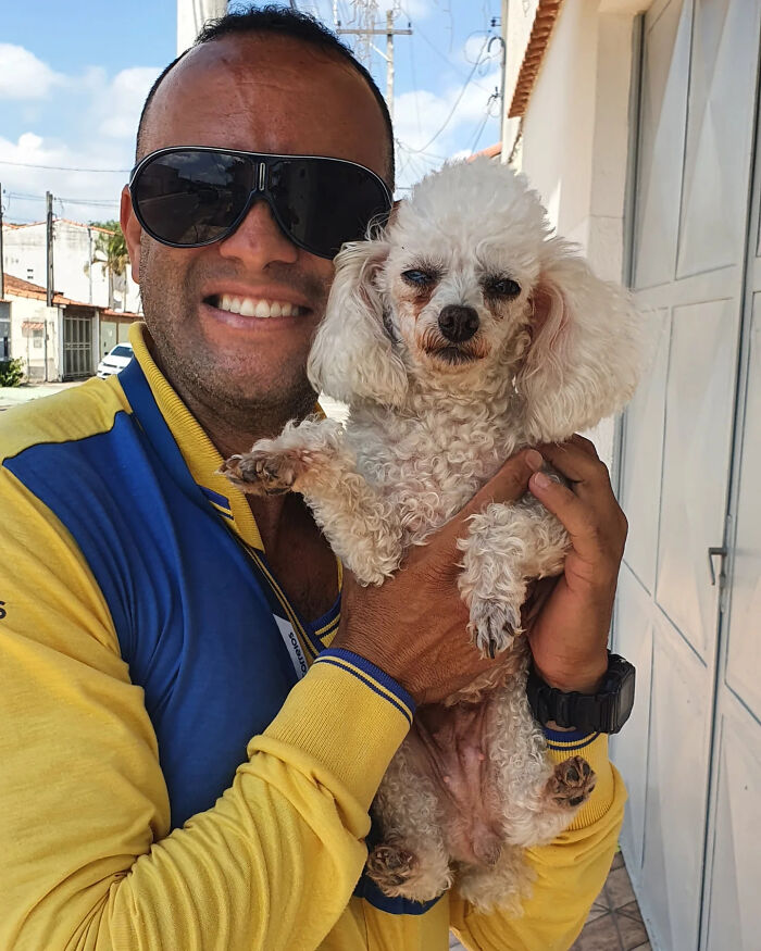Mailman in a yellow and blue jacket holding a small curly white dog, showing their friendly bond on his route.