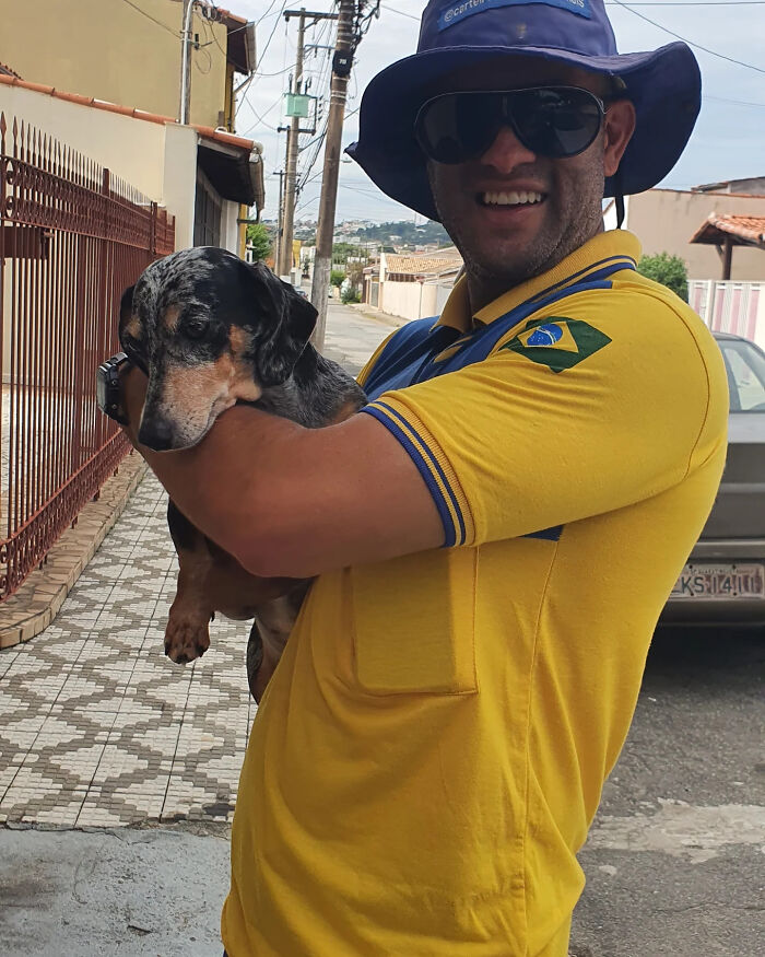 Mailman wearing a yellow uniform and hat smiling while holding a small dog on his delivery route.