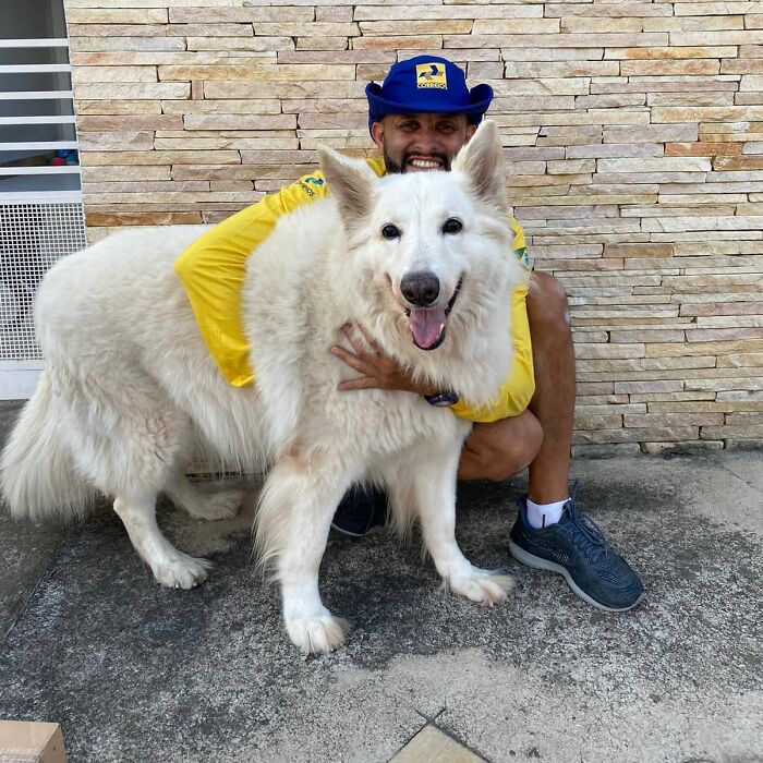 Mailman in uniform kneeling and hugging a large white dog, showing a friendly bond on his delivery route.