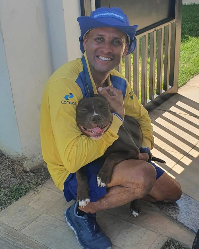 Mailman wearing yellow uniform smiling and hugging a happy dog on his delivery route outdoors.