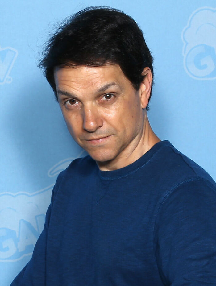 Ralph Macchio in a casual blue shirt posing against a light blue backdrop for bio and career highlights.