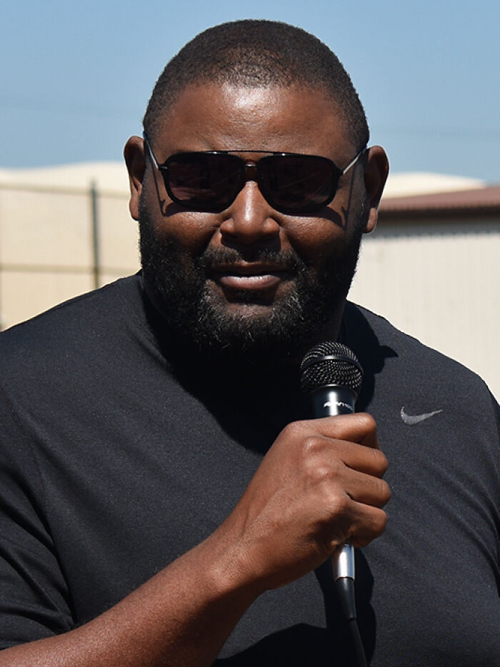 Orlando Pace speaking outdoors, wearing sunglasses and a black Nike shirt, holding a microphone during an event.