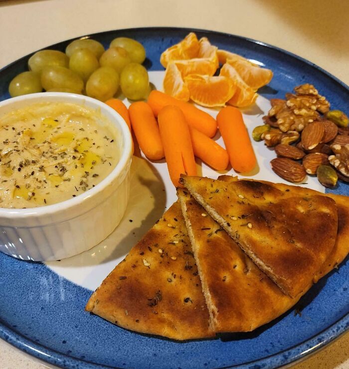 Plate with classic girl dinner combos including hummus, baby carrots, fruit, nuts, and toasted flatbread on a blue plate.