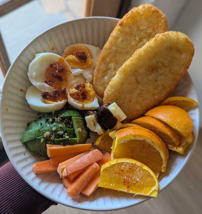 Girl dinner combo with boiled eggs, avocado, carrot sticks, sliced oranges, cheese, olives, and hash browns on a plate.