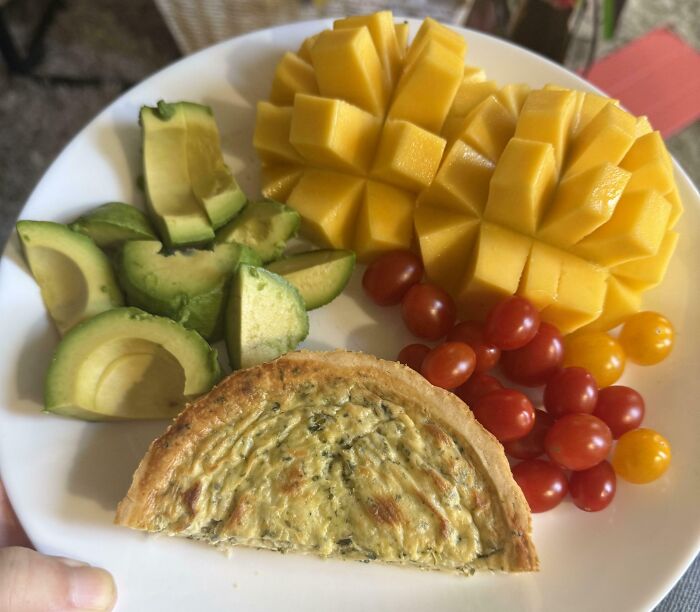 Plate with avocado slices, mango chunks, cherry tomatoes, and a small spinach quiche in a girl dinner combo.