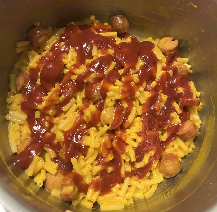 Macaroni and cheese with sliced sausage and ketchup drizzle as a popular girl dinner combo in a metal bowl.