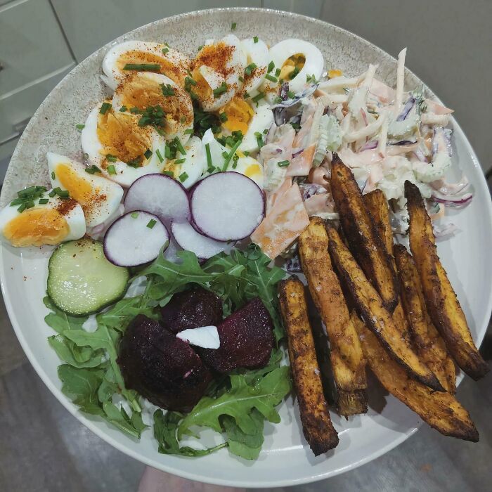 A colorful girl dinner plate with boiled eggs, radishes, arugula, beets, coleslaw, cucumber, and seasoned sweet potato fries.