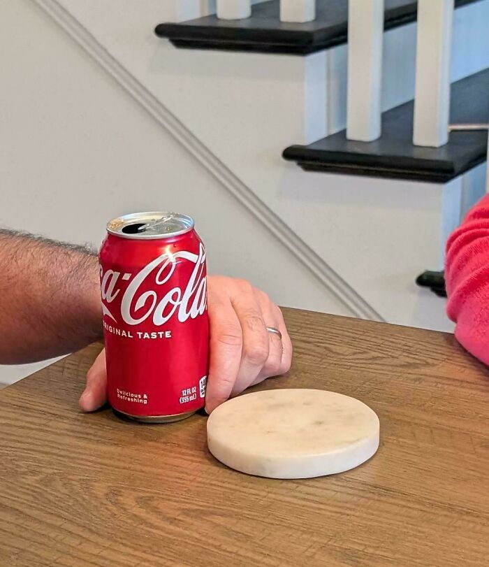Hand holding a Coca-Cola can placed on a wooden table next to an empty coaster showing infuriated family moments.
