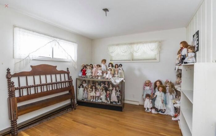 Room in a Zillow home filled with antique dolls, wooden bench, hardwood floors, and white walls with windows and curtains.
