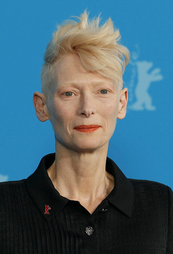 Tilda Swinton with short blond hair and red lipstick, wearing a black collared shirt at a blue backdrop event.