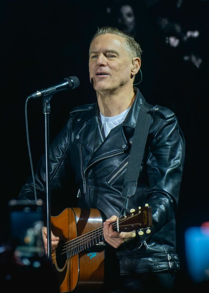 Bryan Adams performing live on stage wearing a black leather jacket while playing an acoustic guitar.