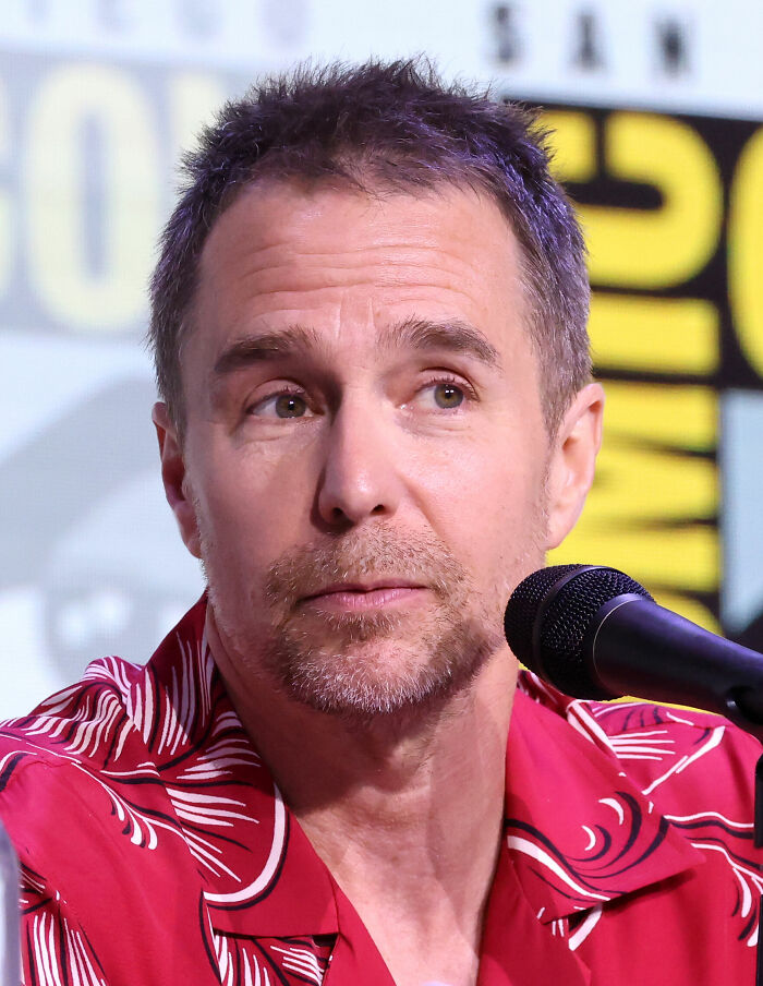 Sam Rockwell speaking at a panel event, wearing a red patterned shirt with a microphone in front of him.
