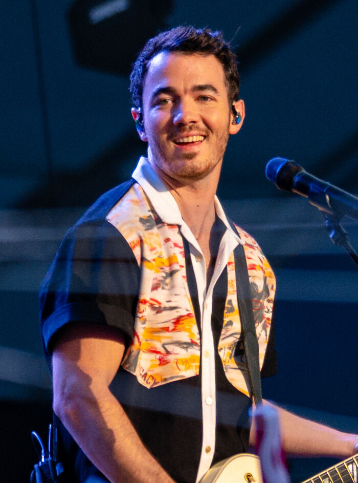 Kevin Jonas smiling on stage during a live music performance wearing a patterned shirt and playing guitar.