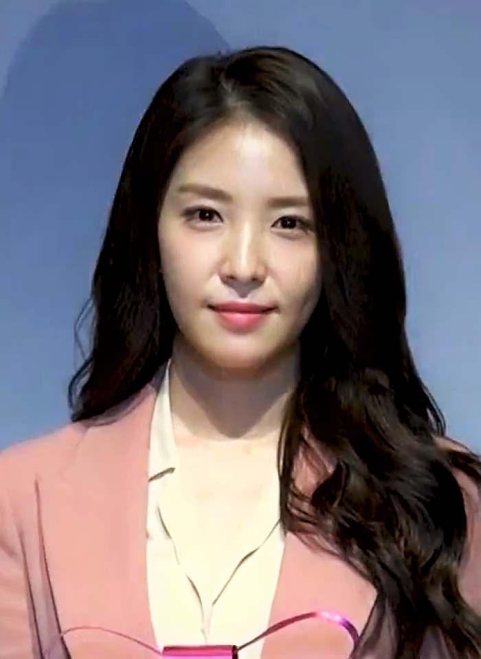 BoA smiling at a public event, wearing a light pink blazer and white blouse with long wavy hair.