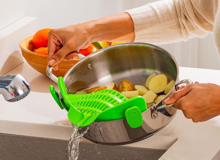 Person using a green kitchen strainer attached to a pot to drain water from peeled potatoes and vegetables.