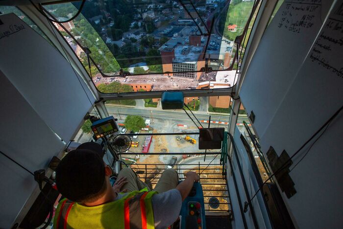 Construction worker operating machinery inside a crane cabin, showing the intricate controls and urban view below.