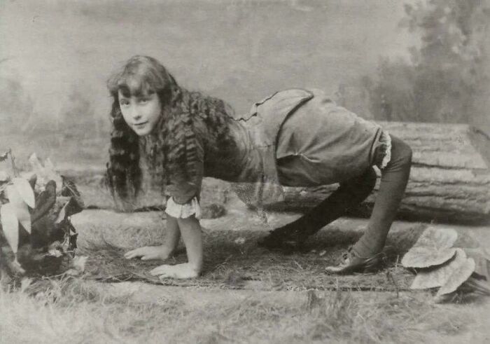 Young woman with long curly hair in Victorian-Edwardian pictures performing a pose on a rustic outdoor setting.
