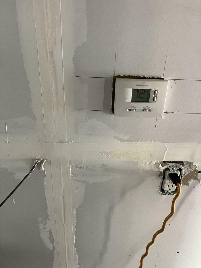 Thermostat installed unevenly with rough drywall work and an exposed electrical outlet, showing people not even trying.