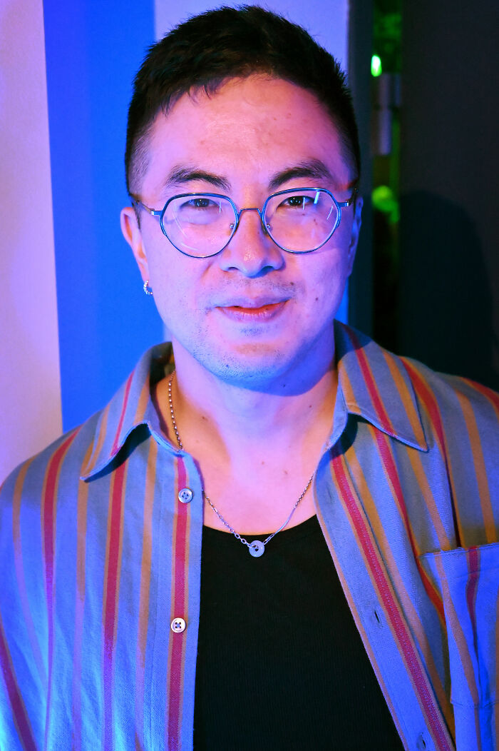 Bowen Yang wearing glasses and a striped shirt, smiling under blue lighting in a casual indoor setting.