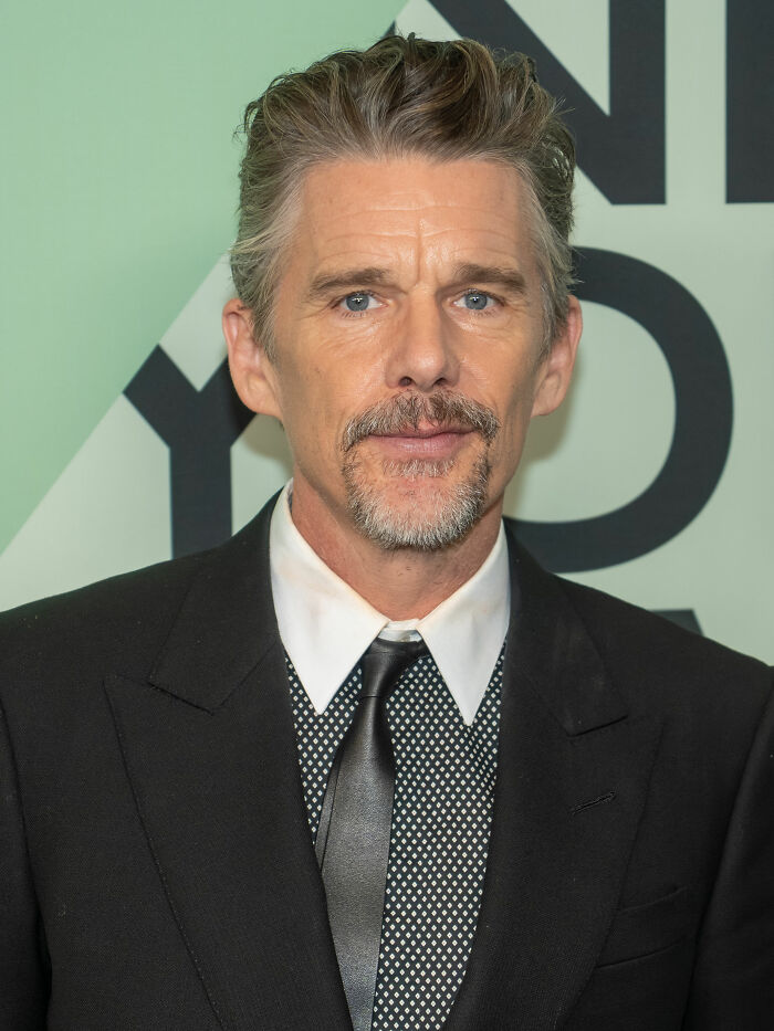 Ethan Hawke at an event wearing a black suit and tie, highlighting his bio and career achievements.