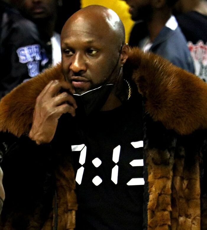 Lamar Odom wearing a fur coat and adjusting his face mask in a crowded indoor setting.