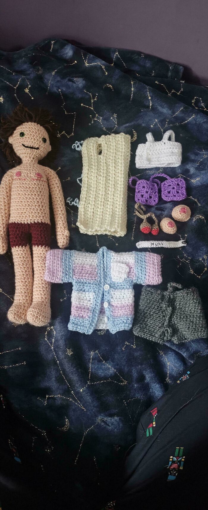 Crocheted doll with neatly arranged handmade clothes and accessories on a dark patterned fabric, showcasing order and craft.