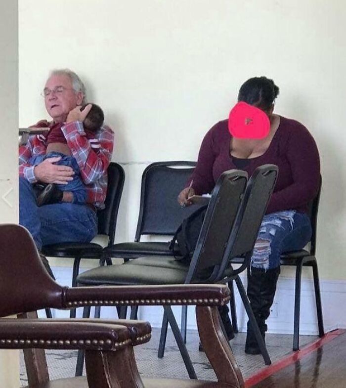 Older man showing faith in humanity restored by gently holding a sleeping child while a woman writes nearby in a waiting room.