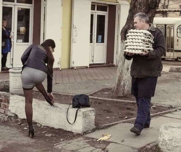 Woman in high heels adjusting shoe near a bench as man carries a stack of frozen frames of egg cartons outdoors.