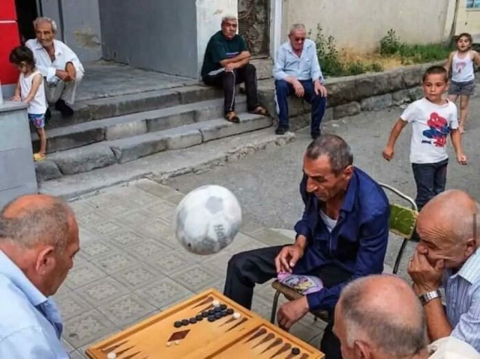 Men playing backgammon outdoors with a soccer ball frozen midair, capturing a moment without showing the aftermath.