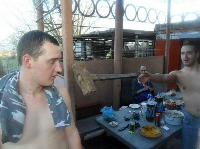 Two shirtless men outdoors frozen in a moment with a large hammer mid-air, capturing a frozen frame of suspense.