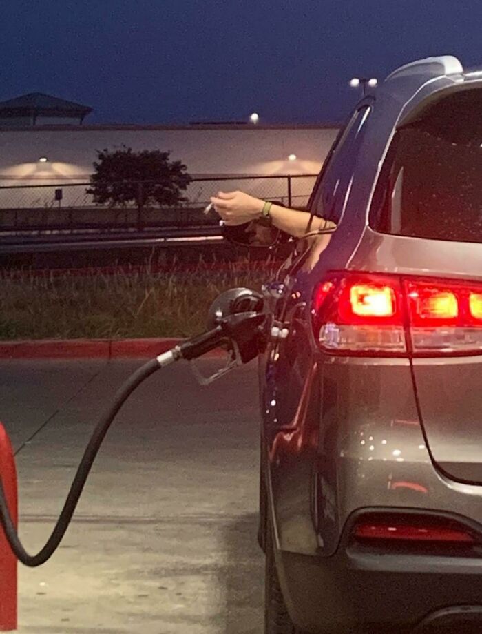 Car at gas station pumping fuel while driver holds a cigarette outside the window in a frozen frame moment at dusk