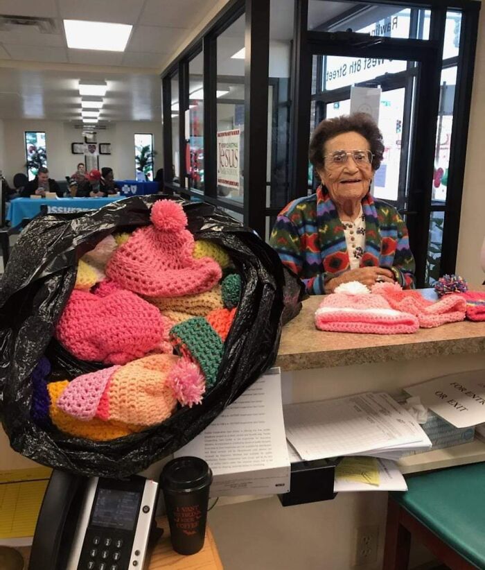 Elderly woman smiling behind a counter filled with colorful hand-knitted hats, showcasing faith in humanity restored.