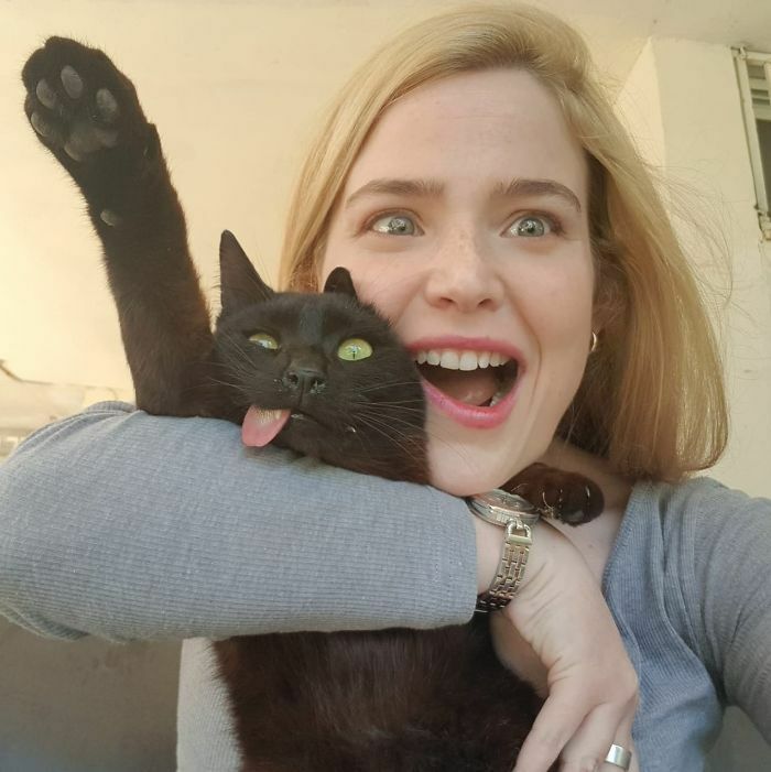 Woman holding a black cat with its tongue out and paw raised, showcasing animals who can take better selfies than you.