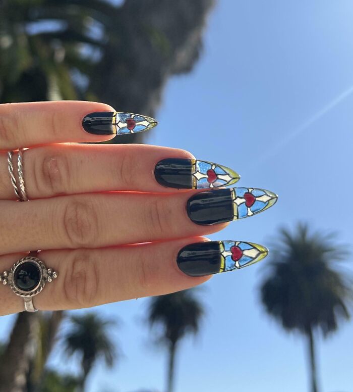 Hand with creative nail art featuring stained glass heart designs against blue sky and palm trees, showcasing artistic projects.