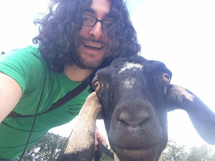 Man with curly hair and glasses taking a close-up selfie with a black and white goat outdoors, showcasing animals selfie skills.