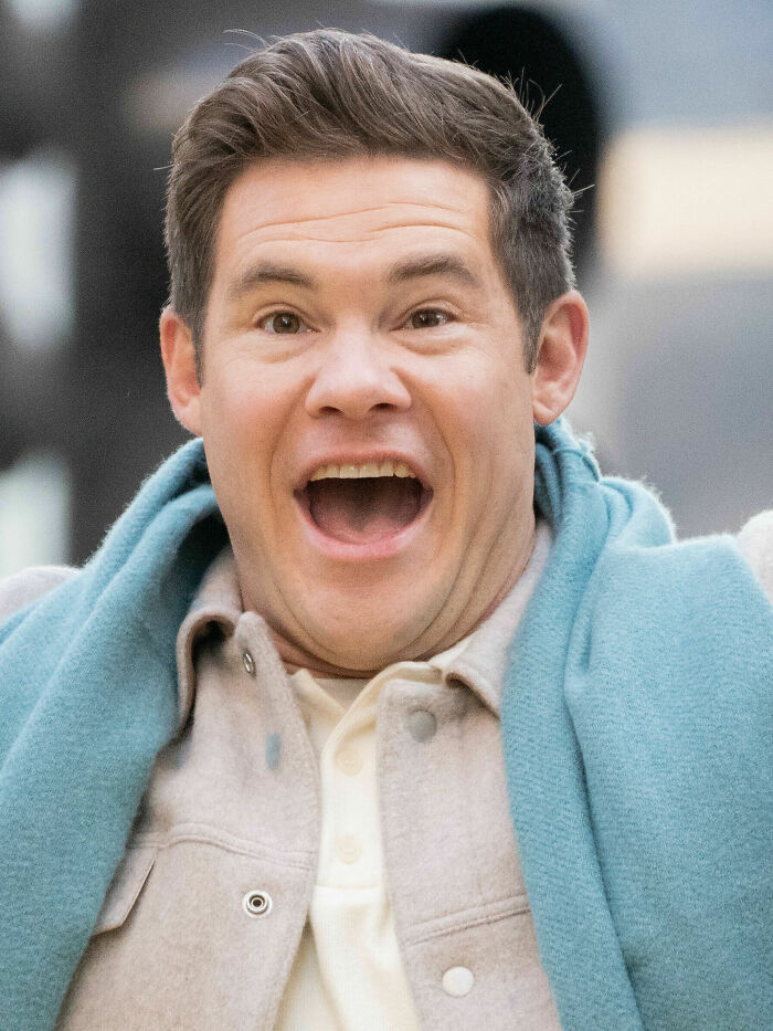Adam DeVine smiling widely outdoors, wearing a light jacket and a blue scarf in a casual setting.